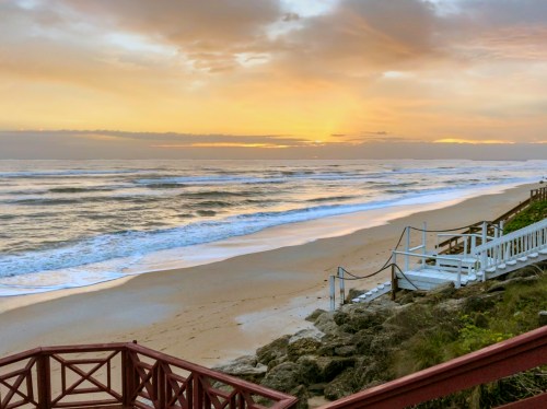 A serene beach at sunset, with gentle waves lapping the shore and a wooden walkway leading to the water.