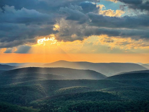 A scenic view of rolling green hills under a dramatic sky with sunlight breaking through clouds at sunset.