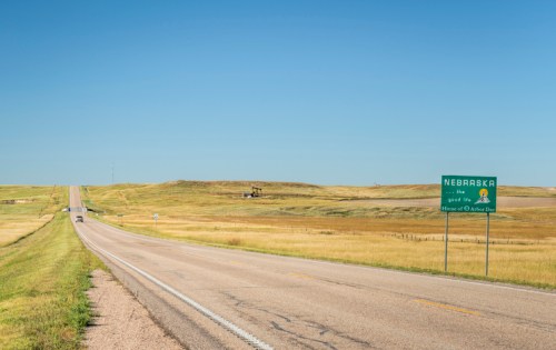 A wide, open road leads into Nebraska, with a welcome sign and rolling green hills under a clear blue sky.