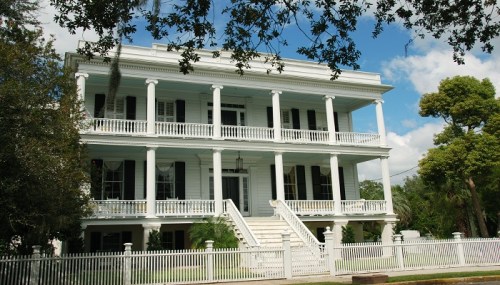 A large, historic white house with columns, surrounded by trees and a white picket fence under a blue sky.