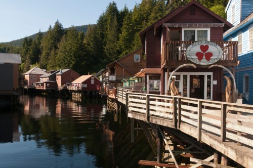 A quaint waterfront scene featuring colorful buildings and a wooden walkway along a calm river surrounded by trees.