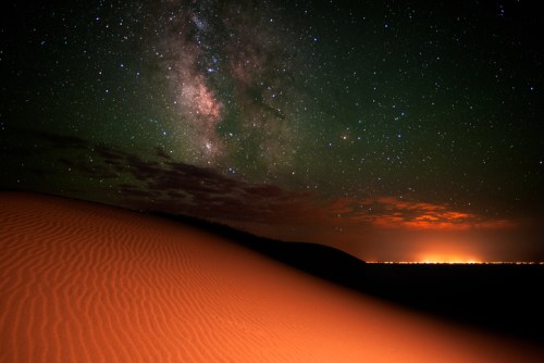 A desert landscape at night, featuring a starry sky with the Milky Way and glowing city lights on the horizon.