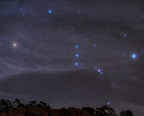 A starry night sky filled with constellations, including Orion, against a dark backdrop of clouds and silhouettes of trees.