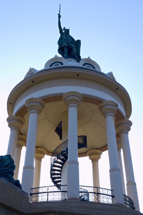 A tall statue of a warrior atop a circular pavilion with columns and a spiral staircase. Clear blue sky in the background.