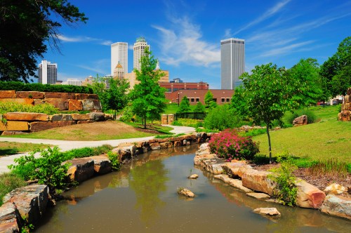A serene park scene featuring a winding stream, lush greenery, and city skyscrapers under a bright blue sky.