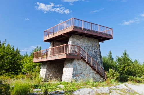 A stone observation tower with wooden railings, surrounded by greenery and under a clear blue sky.