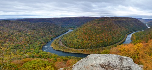 A scenic view of a winding river surrounded by colorful autumn foliage and rolling hills under a cloudy sky.