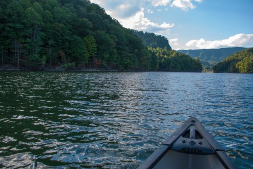 A view from a canoe on a calm lake, surrounded by lush green hills and a clear blue sky.