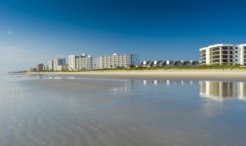 A serene beach scene with calm water reflecting modern buildings under a clear blue sky.