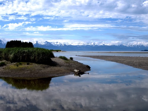 A serene landscape featuring a calm river reflecting mountains and clouds, surrounded by lush greenery and sandy shores.