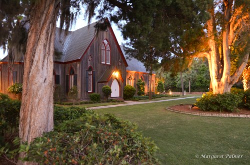 A charming wooden building with arched windows, surrounded by lush greenery and illuminated by soft lights at dusk.