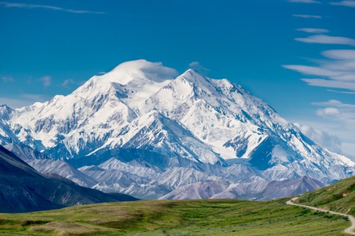 A majestic snow-capped mountain rises against a clear blue sky, surrounded by rolling green hills and rugged terrain.