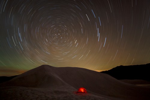 A glowing red tent sits in a desert landscape under a starry night sky with swirling star trails.
