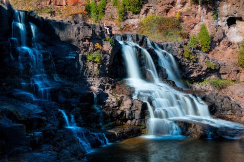A serene waterfall cascades over rocky cliffs, surrounded by lush greenery and vibrant autumn foliage.