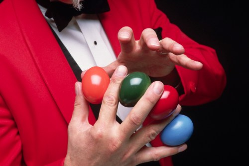 A magician in a red jacket skillfully balances colorful plastic eggs in his hands against a dark background.