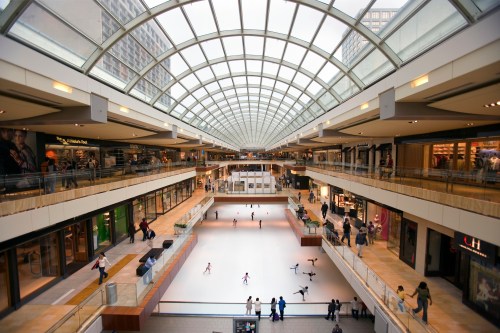 A spacious shopping mall with a glass ceiling, featuring an ice skating rink and shoppers on multiple levels.