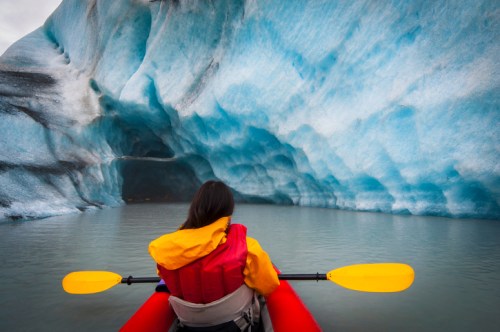A person in a kayak paddles towards a blue glacier cave, surrounded by icy formations and calm water.