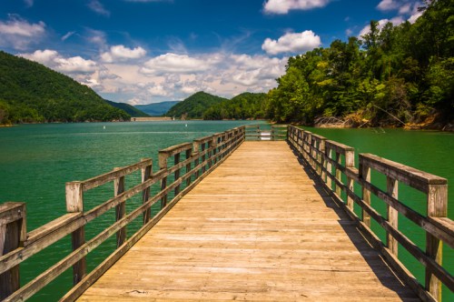 A wooden pier extends into a serene green lake, surrounded by lush mountains and a blue sky with fluffy clouds.