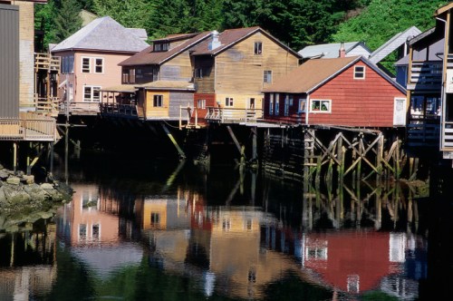 Colorful wooden houses on stilts reflect in calm water, surrounded by lush greenery.