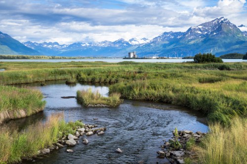 A serene landscape featuring a winding stream, lush green fields, and majestic mountains under a blue sky.