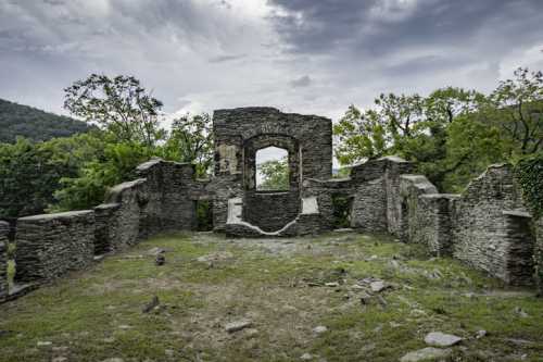 Ruins of a stone structure surrounded by greenery, with a cloudy sky overhead.