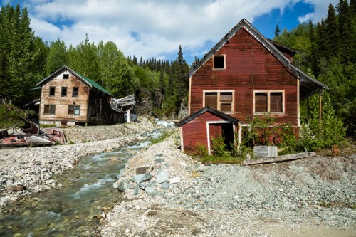 Abandoned red and wooden buildings beside a rocky stream, surrounded by lush green trees and a blue sky.
