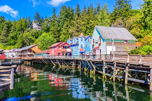 Colorful houses line a wooden pier beside calm waters, surrounded by lush green trees and a clear blue sky.