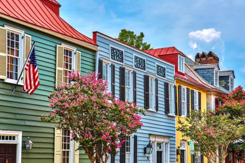 Colorful historic houses with vibrant flowers and an American flag, set against a bright blue sky.