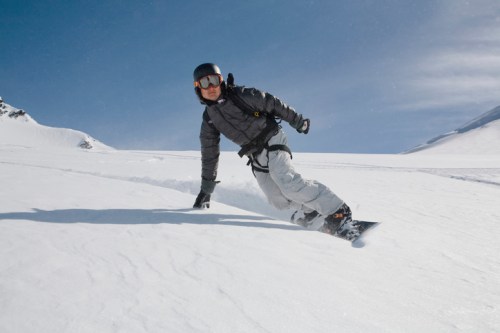 A snowboarder in a black helmet and gray jacket carves through fresh snow on a sunny mountain slope.