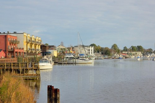 A calm waterfront scene with boats docked, colorful buildings lining the shore, and a cloudy sky above.