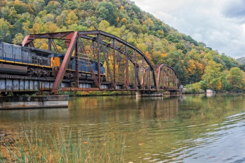 A train crosses a rusted bridge over a calm river, surrounded by colorful autumn foliage on the hills.