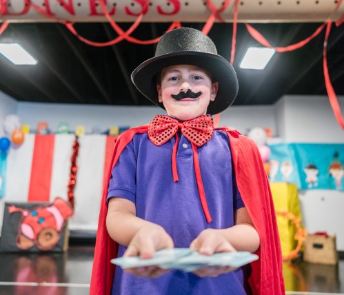 A child in a top hat and cape holds playing cards, smiling in a festive, decorated room.
