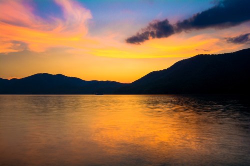 A serene sunset over a calm lake, with vibrant colors reflecting on the water and mountains in the background.