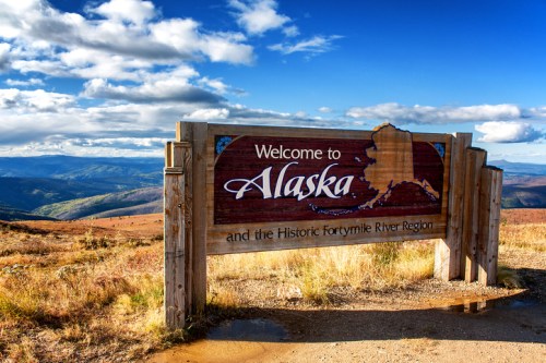 A wooden sign welcomes visitors to Alaska, with scenic mountains and a blue sky in the background.