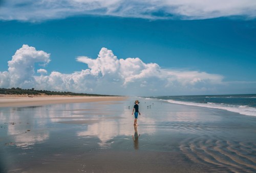 A child walks along a sandy beach, reflecting clouds and blue sky in the water, with seagulls in the distance.