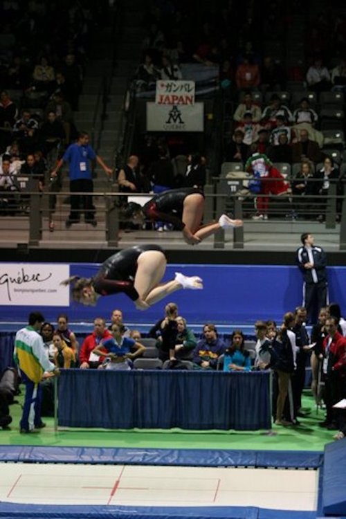 Two gymnasts performing flips in mid-air during a competition, with spectators and officials in the background.