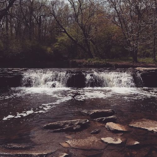 A serene waterfall cascades over rocks, surrounded by lush trees and a calm river in a peaceful natural setting.
