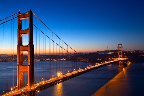 Golden Gate Bridge illuminated at dusk, with a clear sky and city lights reflecting on the water.