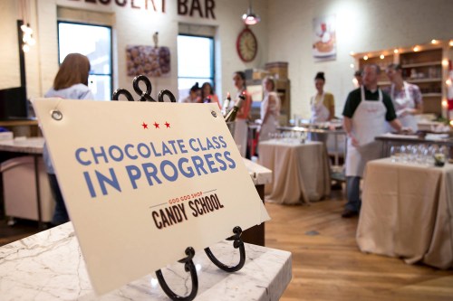 Sign reading "Chocolate Class in Progress" in a cozy kitchen setting with people preparing for a candy-making class.