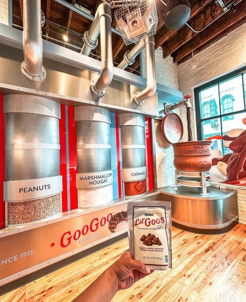 A hand holds a bag of Lil' Goos in front of a colorful display of ingredients at a candy shop.