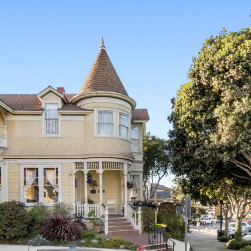 A charming yellow Victorian house with a turret, surrounded by greenery and trees on a sunny day.