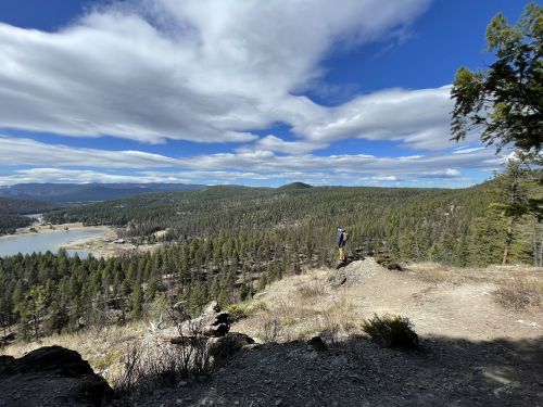 A hiker stands on a rocky outcrop overlooking a lush valley and lake, with dramatic clouds in the blue sky above.