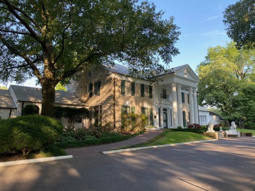A large, elegant house with white columns, surrounded by trees and greenery, on a sunny day.