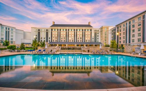 A hotel with a large pool reflects a colorful sunset sky, surrounded by lounge chairs and landscaped greenery.