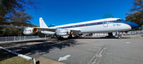 A vintage airplane parked on a tarmac under a clear blue sky, surrounded by trees and a fence.