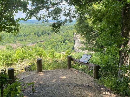 A scenic overlook surrounded by trees, featuring a sign and a view of a lush green valley below.
