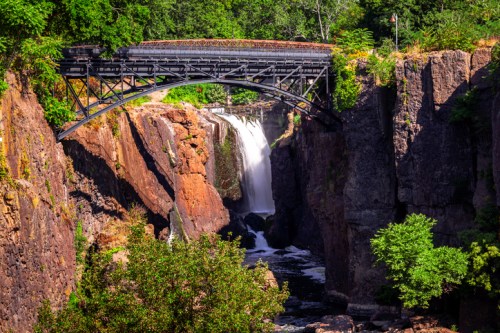 A scenic view of a waterfall cascading between rocky cliffs, with a bridge arching above lush greenery.