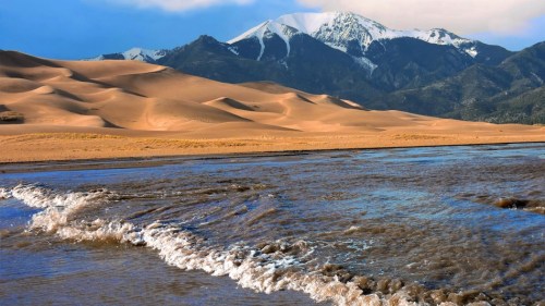 A scenic view of sandy dunes in the foreground with a snow-capped mountain in the background and a river in the foreground.