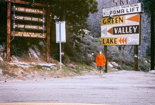A person in an orange coat stands at a road intersection with various directional signs in a mountainous area.