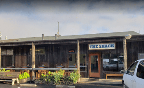 A rustic building with a sign reading "The Shack" and an "Open" neon sign, featuring outdoor seating.